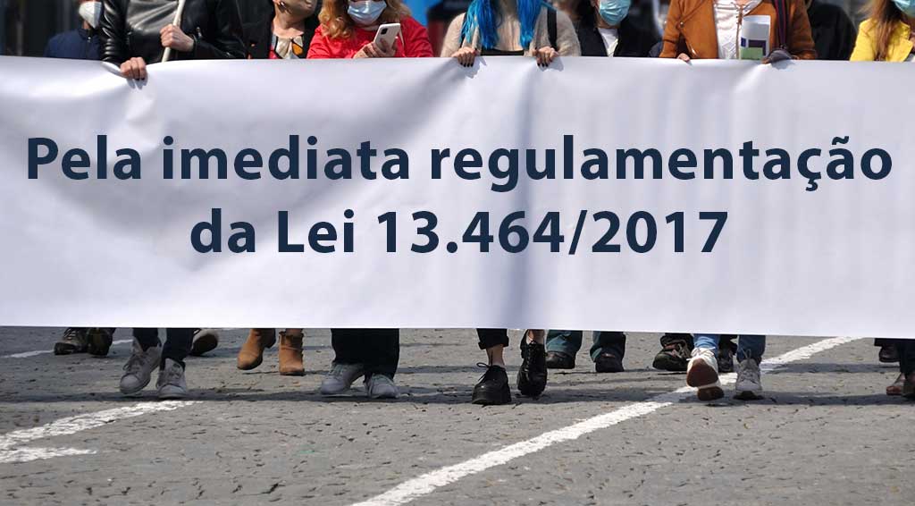 Pessoas segurando um cartaz branco com o dizer "Pela imediata regulamentação da Lei 13.464/2017"